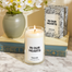 A close up of the In Our Hearts candle on a marble surface. Behind the candle is a book that has a plant on top and the packaging for the candle.
