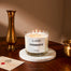 A lifestyle image of the 3-Wick Hogwarts candle displayed on a round white marble coaster. The coaster sits on a dark wood table with gold props also on it.