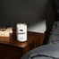 A bedside shot of the Massachusetts Candle on a deep glossy wood side table. One can see a cream book in the corner and the silky gray sheets of the bed.