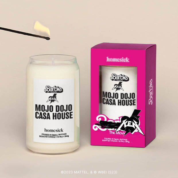 A lit Mojo Dojo Case House Homesick candle displayed next to its boxed packaging on a dark cream background.