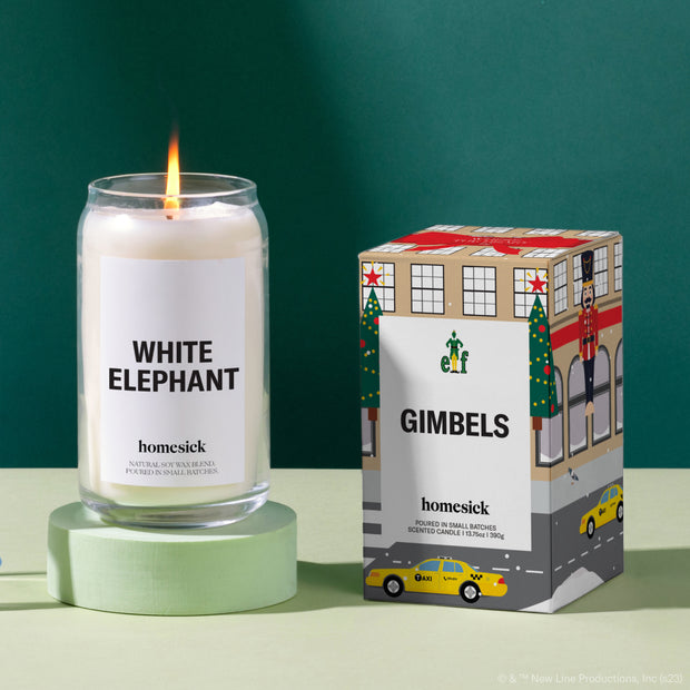 A lit White Elephant Candle on a mint green pedestal. Next to it is the boxed Gimbels candle from the Elf collection. Both are on a mint green surface with a dark green background.