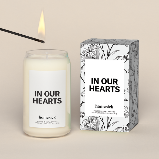 A lit In Our Hearts Homesick candle displayed next to its boxed packaging on a dark cream background.
