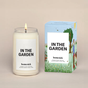 A lit In The Garden Homesick candle displayed next to its boxed packaging on a dark cream background.