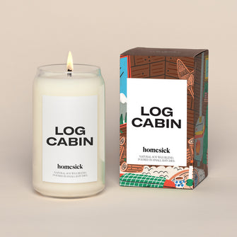 A lit Log Cabin Homesick candle displayed next to its boxed packaging on a dark cream background.