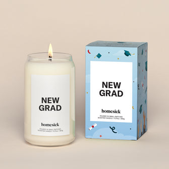 A lit New Grad Homesick candle displayed next to its boxed packaging on a dark cream background.
