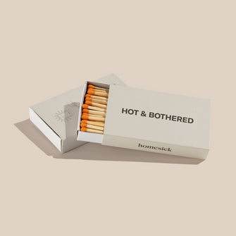 A box of the Hot and Bothered Homesick candles shot on a dark cream background. The top box is slid slightly open to display the tips of the matches.