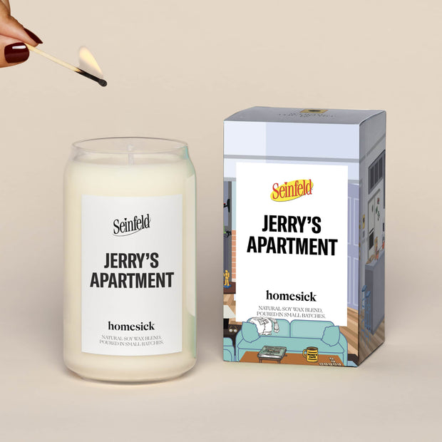 A lit Jerry's Apartment Homesick candle displayed next to its boxed packaging on a dark cream background.