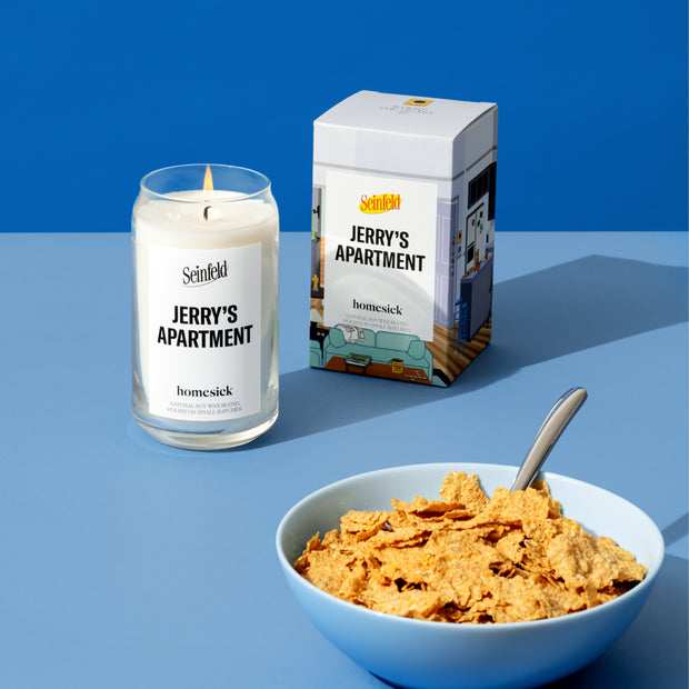 A stylized studio shot of the Jerry's Apartment candle from Homesicks Seinfield collection. There is a bowl of cereal in the lower right corner of the image. In the back left and middle is the Jerry's Apartment candle and packaging. They are shot on a light blue surface with a darker blue background.