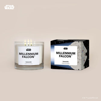 A lit 3-Wick Millennium Falcon Homesick candle displayed next to its boxed packaging on a dark cream background.