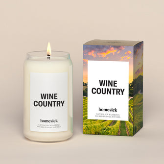 A lit Wine Country Homesick candle displayed next to its boxed packaging on a dark cream background.