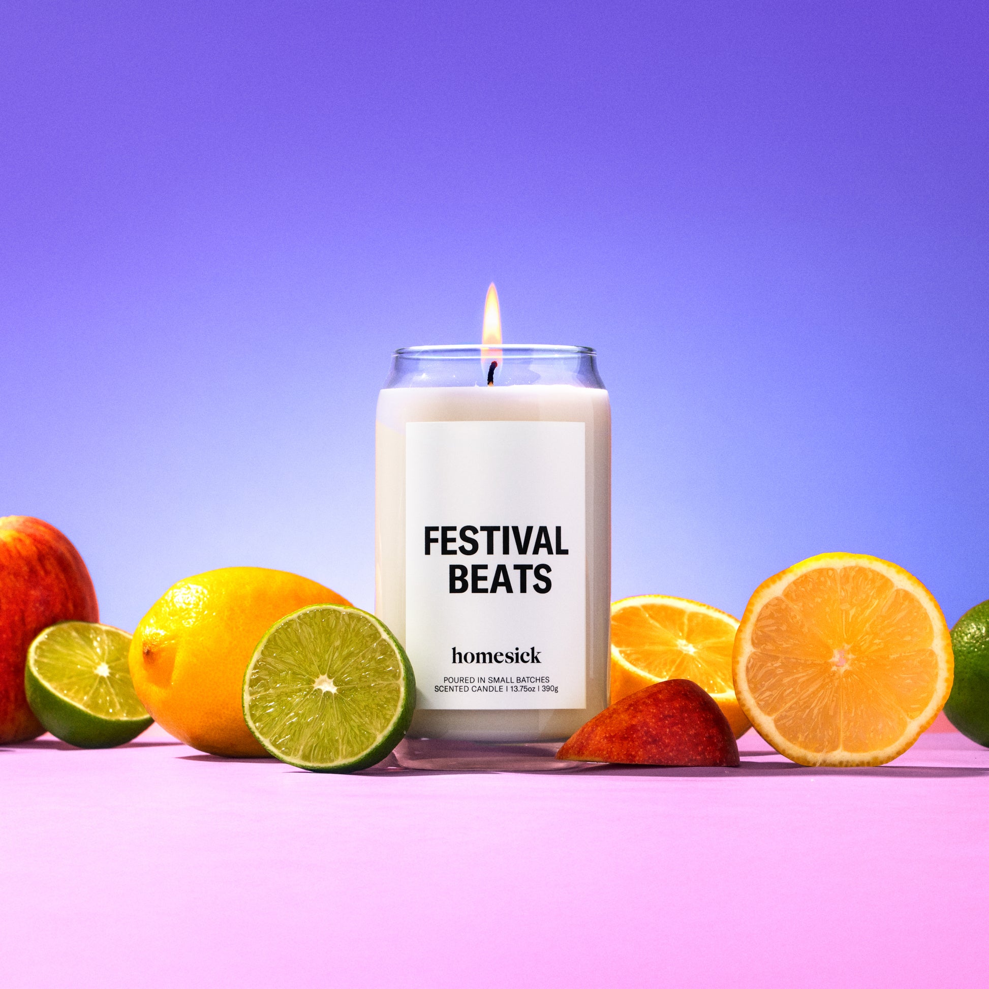 A lit Festival Beats Homesick candle on a colorful background surrounded by various fruits
