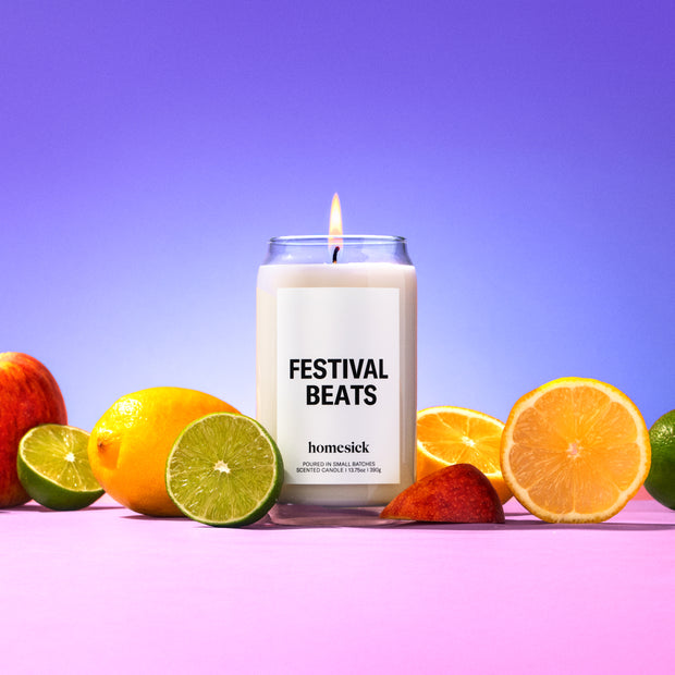 A lit Festival Beats Homesick candle on a colorful background surrounded by various fruits