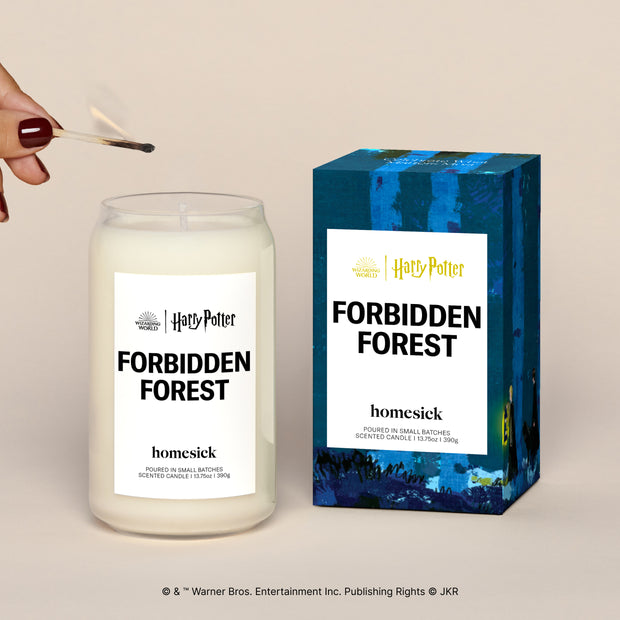 A lit Forbidden Forest Homesick candle displayed next to its boxed packaging on a dark cream background.