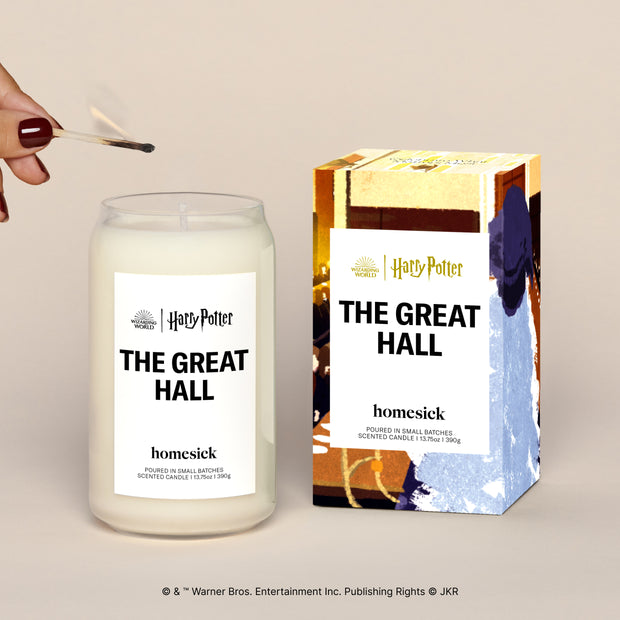 A lit The Great Hall candle displayed next to its boxed packaging on a dark cream background.