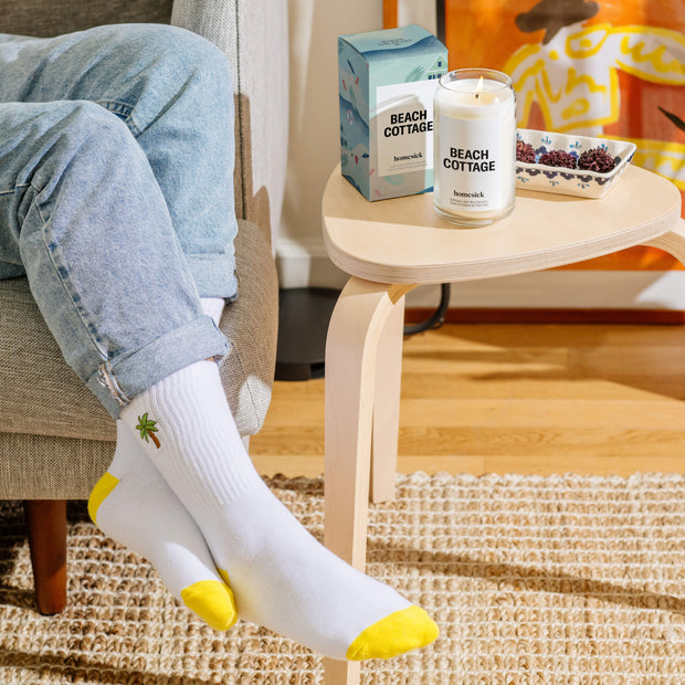 A shot of a model wearing the palm high rise socks with their feet overhanging the couch. Next to their feet is a side table with the beach cottage candle