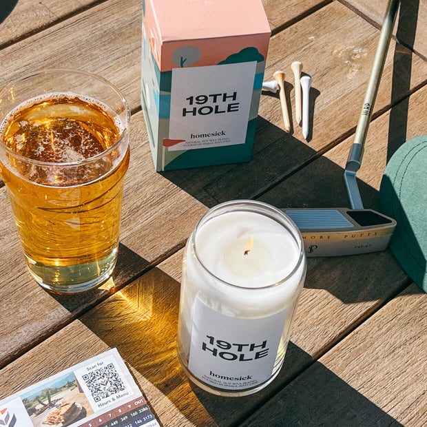 A lifestyle image of the 19th Hole Homesick candle with various golf props surrounding it. 