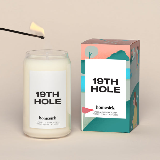 A lit 19th Hole Homesick candle displayed next to its boxed packaging on a dark cream background.