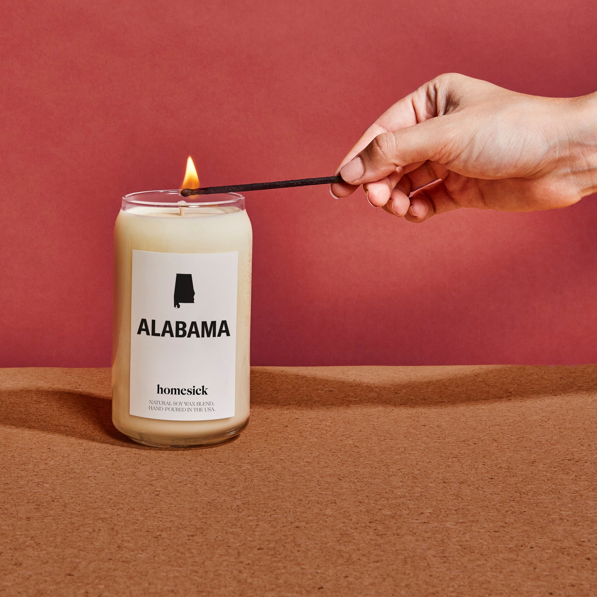 A hand using a match to light the Alabama Homesick candle. The candle is on a cork board surface with a red background.