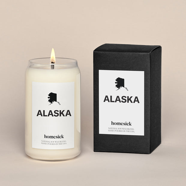 A lit Alaska Homesick candle displayed next to its boxed packaging on a dark cream background.
