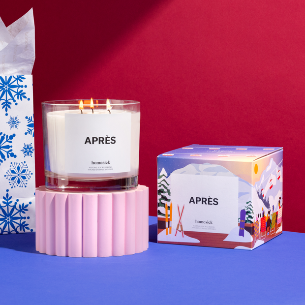 The larger size of the Après candle lit on a pink pedestal next to its boxed packaging. These two items are on a cobalt blue table with a red background.