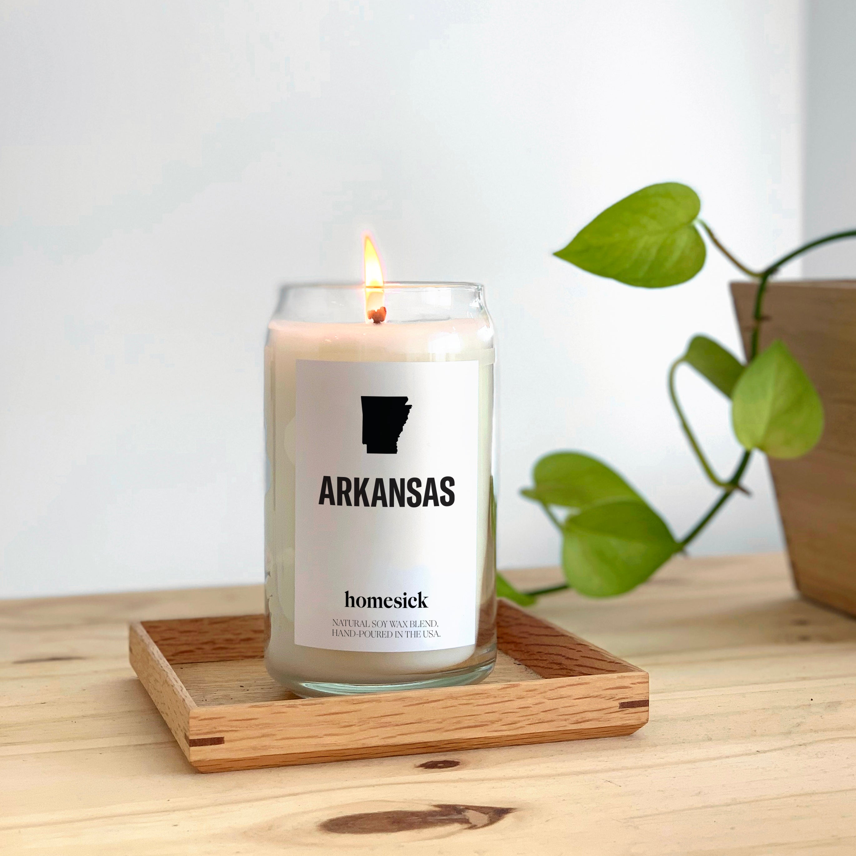 A lit Arkansas candle placed inside a square shallow wooden dish on top of a wooden table. One can see a green plant in the corner.