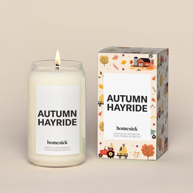 A lit Autumn Hayride Homesick candle displayed next to its boxed packaging on a dark cream background.