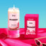 A display of a lit Barbie Dreamhouse candle next to the pink packaged box. Both are displayed on top of a pink table surface with a blue background.