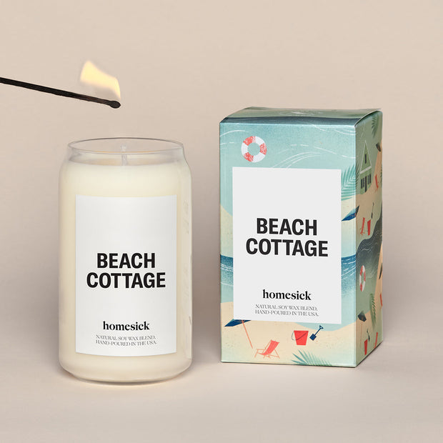 A lit Beach Cottage Homesick candle displayed next to its boxed packaging on a dark cream background.