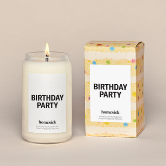 A lit Birthday Party Homesick candle displayed next to its boxed packaging on a dark cream background.
