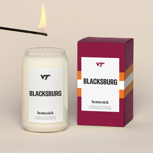 A lit Virginia Tech Blacksburg Homesick candle displayed next to its boxed packaging on a dark cream background.