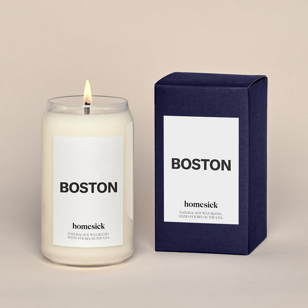 A lit Boston Homesick candle displayed next to its boxed packaging on a dark cream background.