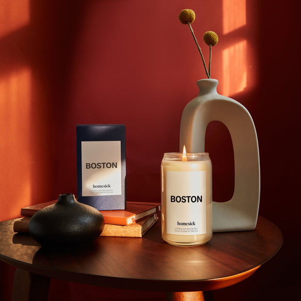 A lifestyle image of the Boston Homesick Candle on a side table with various home decor also on the table. There is a red background and lighting is moody.