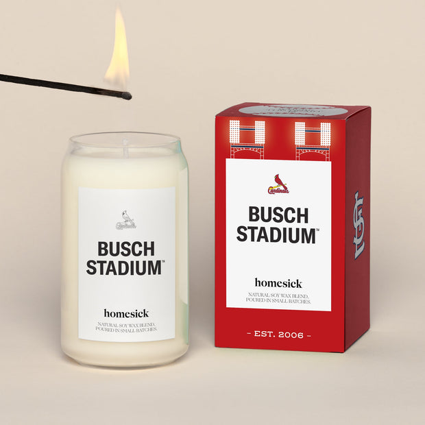 A lit Busch Stadium Homesick candle displayed next to its boxed packaging on a dark cream background.
