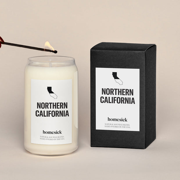 A lit Northern California Homesick candle displayed next to its boxed packaging on a dark cream background.