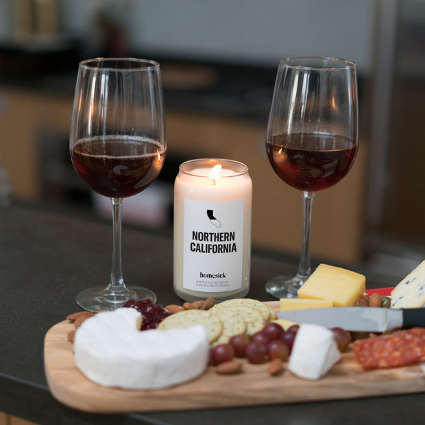 A Northern California candle shot between two glasses of red wine with a charcuterie board in front of those.