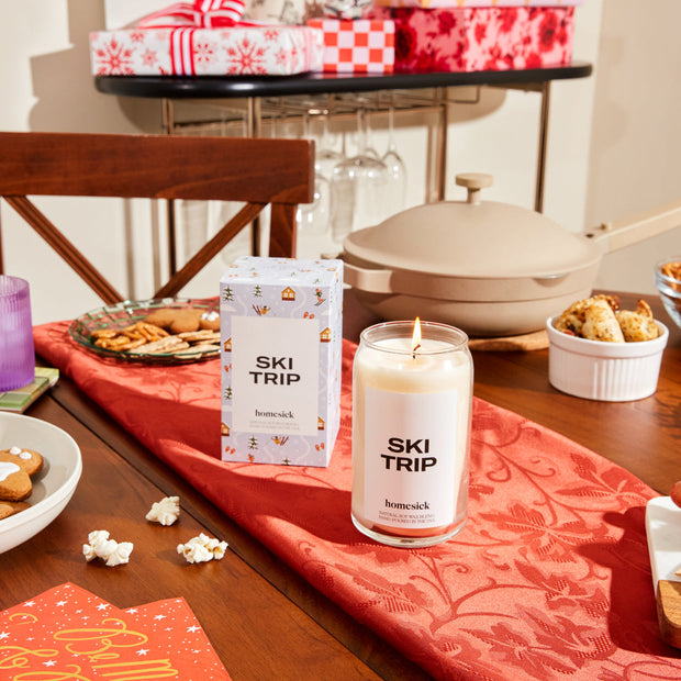 A stylized holiday shot of the Ski Trip candle in the center of a table that is hosting for the holidays. The candle is on top of a red seasonal tablecloth with platters of holiday cookies and presents blurred in the background.