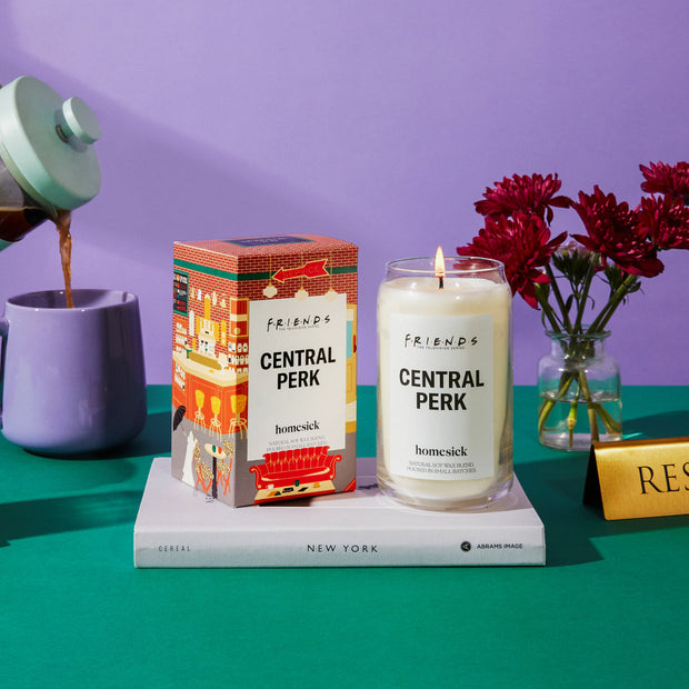 A stylized shot of the Central Park Candle next to its packaging that sit on top of a new york book. The book sits on top of a dark green surface with a purple background.