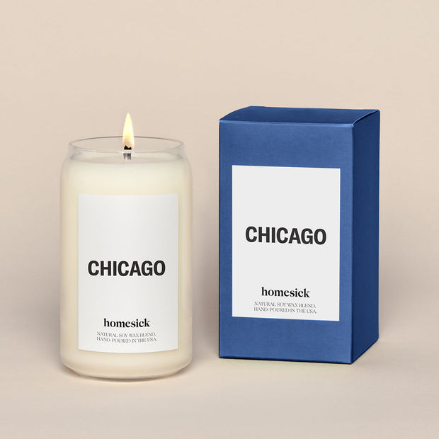 A lit Chicago Homesick candle displayed next to its boxed packaging on a dark cream background.
