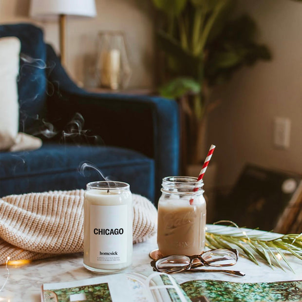 A lifestyle image of a recently blown out Chicago candle on a side table with various items. IN the background, there is a couch, plant and other living room decor.