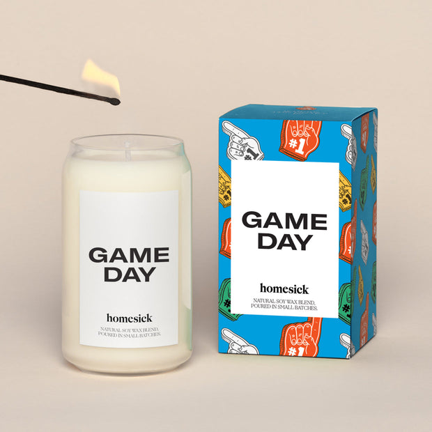 A lit Game day Homesick candle displayed next to its boxed packaging on a dark cream background.