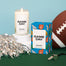 A stylized shot of the Game Day candle and its packaging. Around the candle are football props on a green surface and blue background.