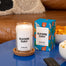 A stylized shot of the game day candle and its packaging on a blue table. The Table has various game day snacks and there is a brown couch in the backgroud.