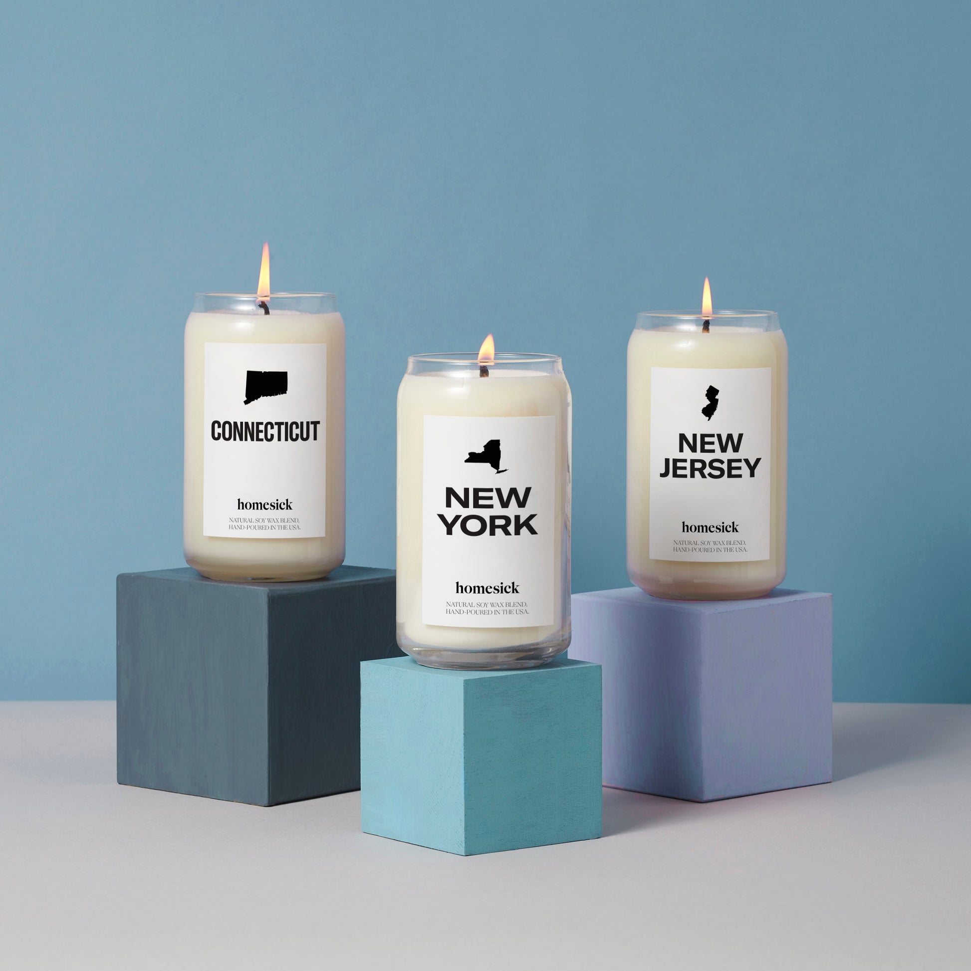 Three homesick candles on top of different colored square pedestals in front of a blue background on a gray surface.