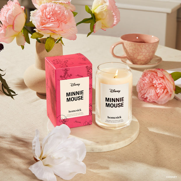 The Minnie Mouse candle and its packaging displayed on top of a circular marble coaster. Surrounding those items are a pink peonies and a cup of tea.