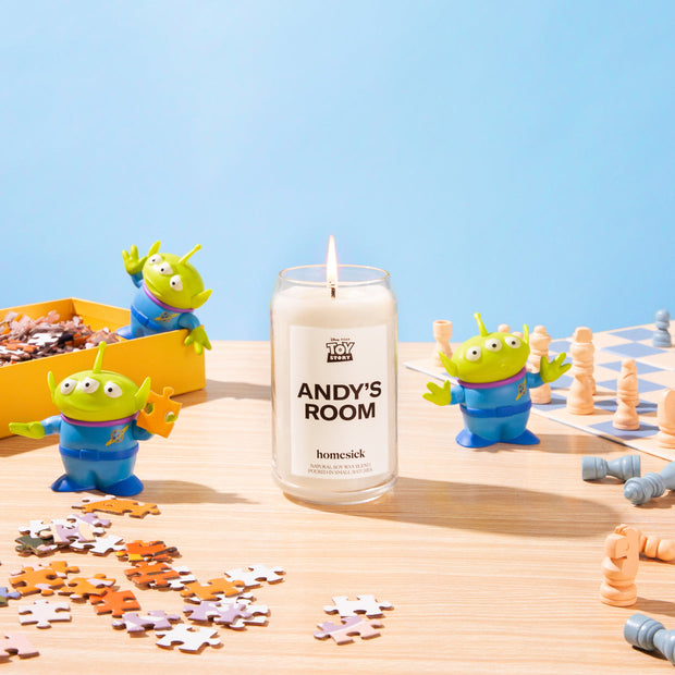 A stylized shot of the Andy's Room candle in the center of a light wood table. There are Toy Story aliens and puzzle pieces around in with a light blue background.