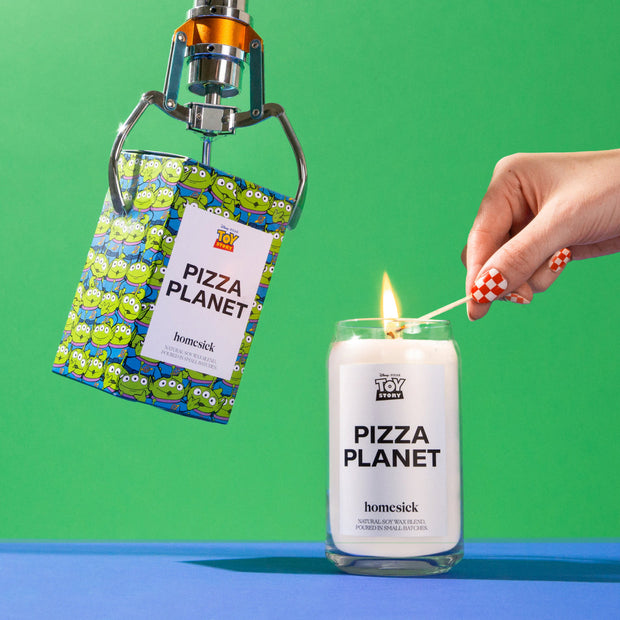 On the left is a claw picking up the Pizza Planet packaging with the Pizza Planet candle next to it. A hand is lighting the candle. The surface is bright blue with a bright green background.