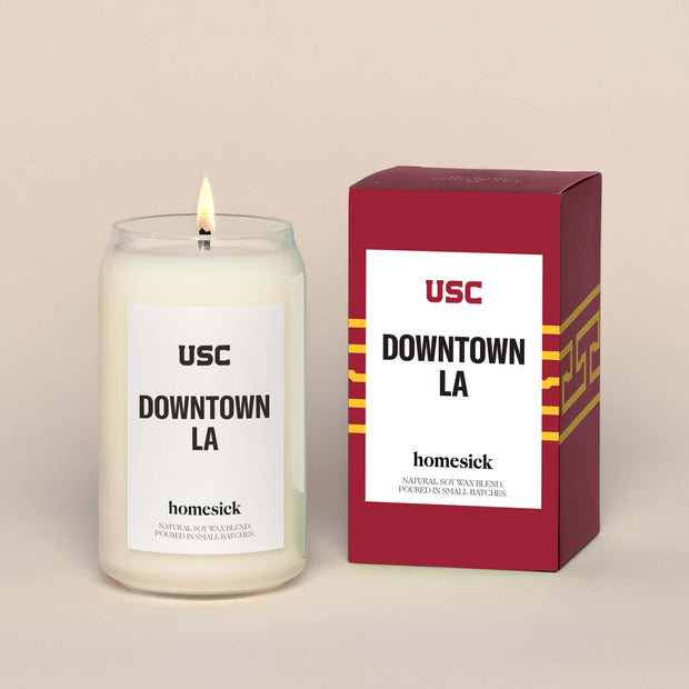 A lit USC Downtown LA  Homesick candle displayed next to its boxed packaging on a dark cream background.
