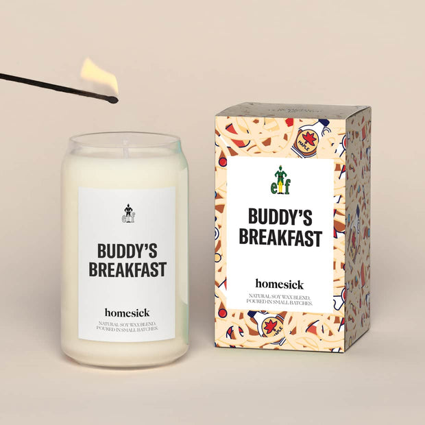 A lit Buddy's Breakfast Homesick candle displayed next to its boxed packaging on a dark cream background.