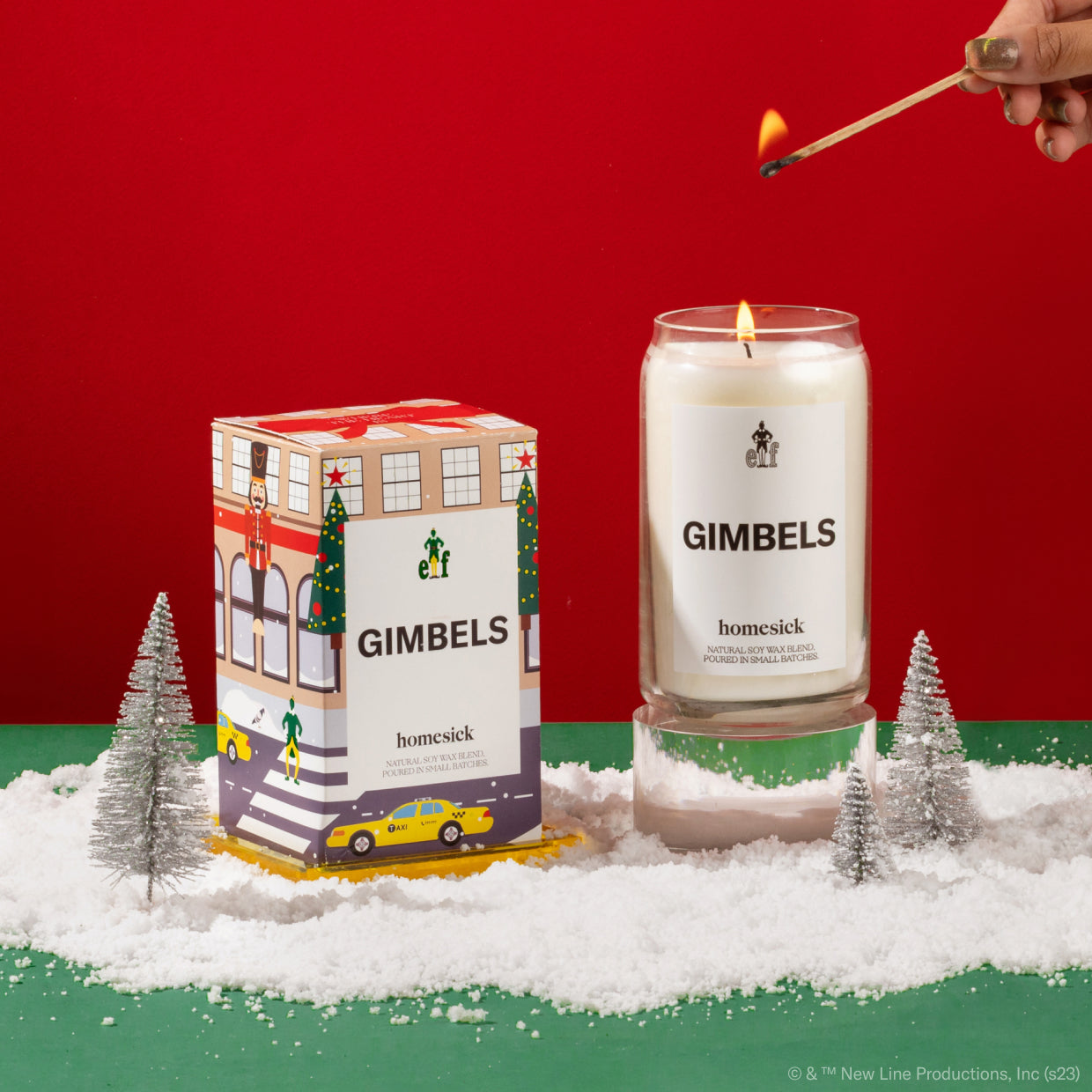 A hand coming in to light the Gimbels candle. Next to the candle is its packaging. Both are shot in a holiday environment on a green surface, with fake snow flakes, and a red background.