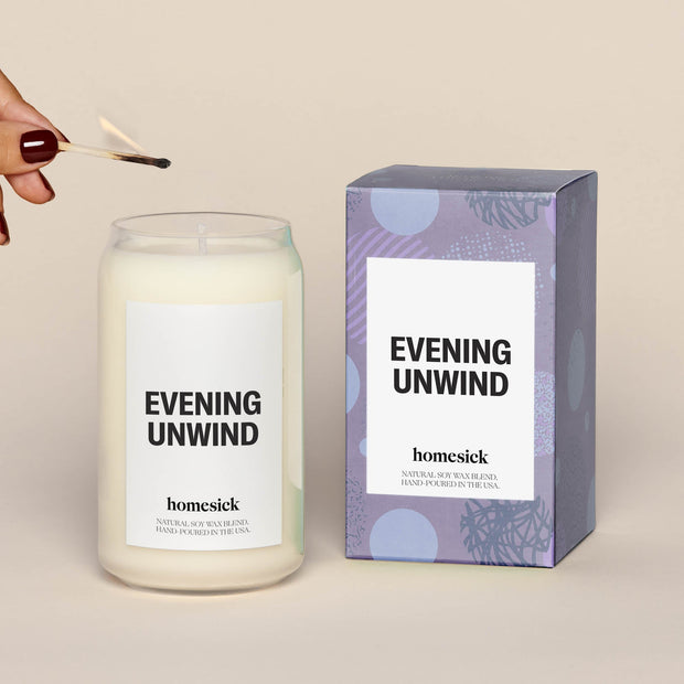 Single evening unwind lit candle next to a purple box, which the candle comes in. 